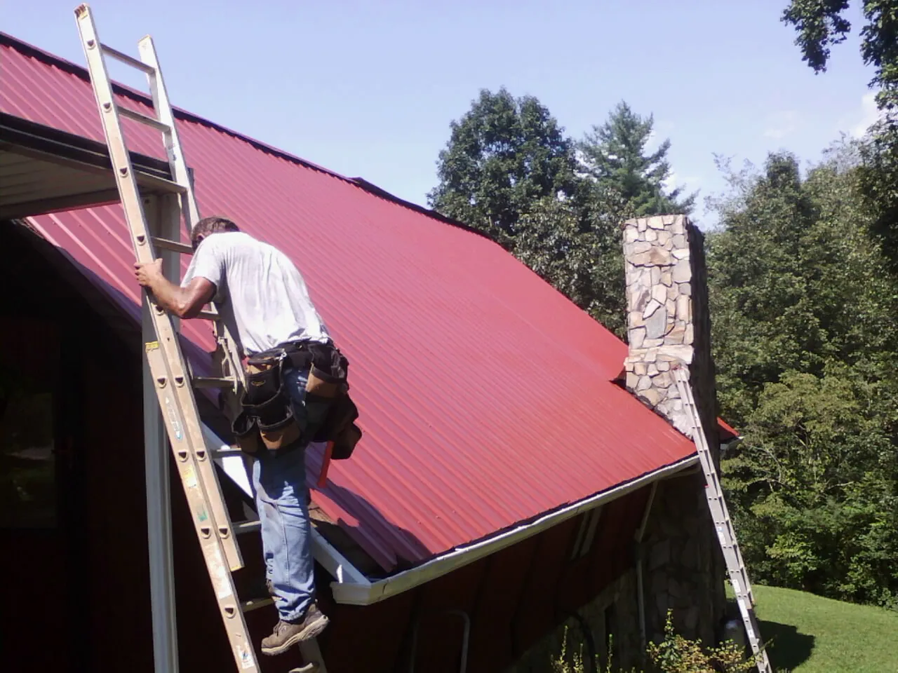 Roofer on a ladder beside a red metal roof and stone chimney — Soffit & Fascia Repair by Roofing Brothers in San Bernardino